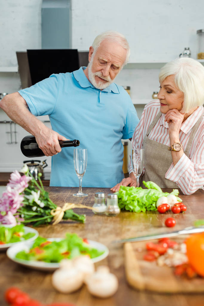 Selective focus of senior couple pouring champagne by bouquet and vegetables on kitchen table - Photo, Image