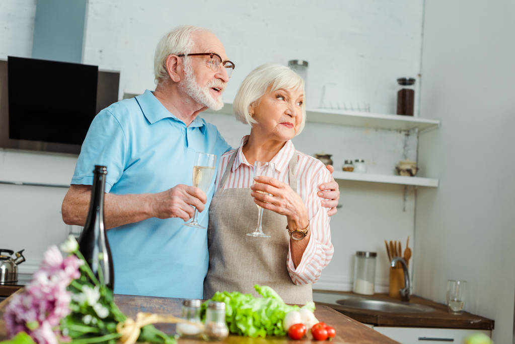 Foco seletivo do casal sênior com copos de champanhe olhando para longe ao lado de buquê e legumes na mesa da cozinha
  - Foto, Imagem