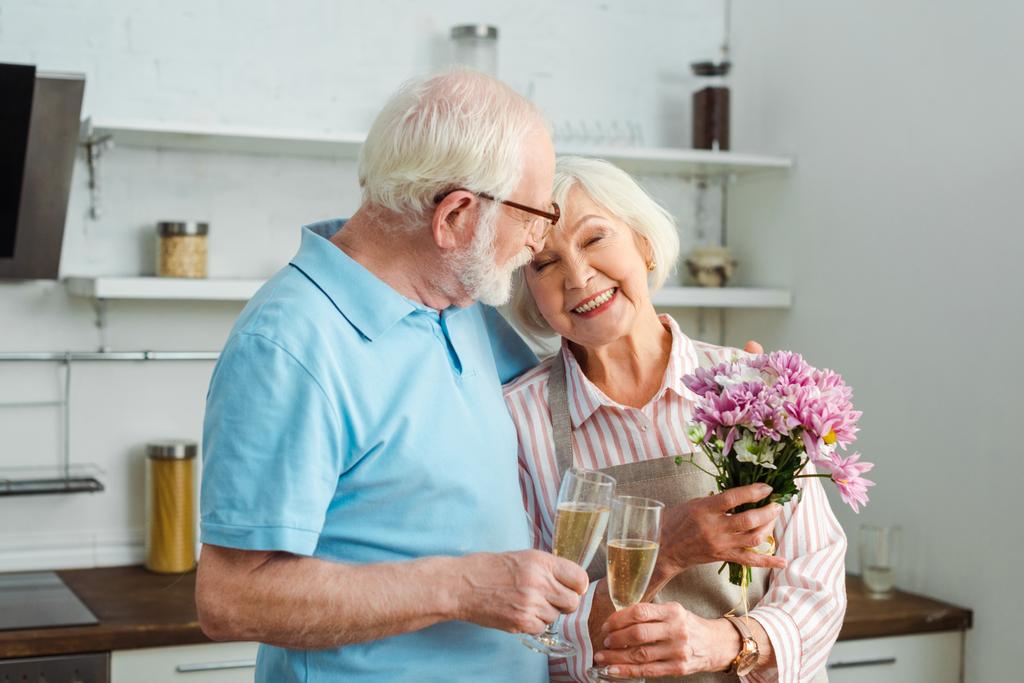 Χαμογελαστή ηλικιωμένη γυναίκα που κρατάει μπουκέτο και πίνει σαμπάνια με τον άντρα της στην κουζίνα. - Φωτογραφία, εικόνα