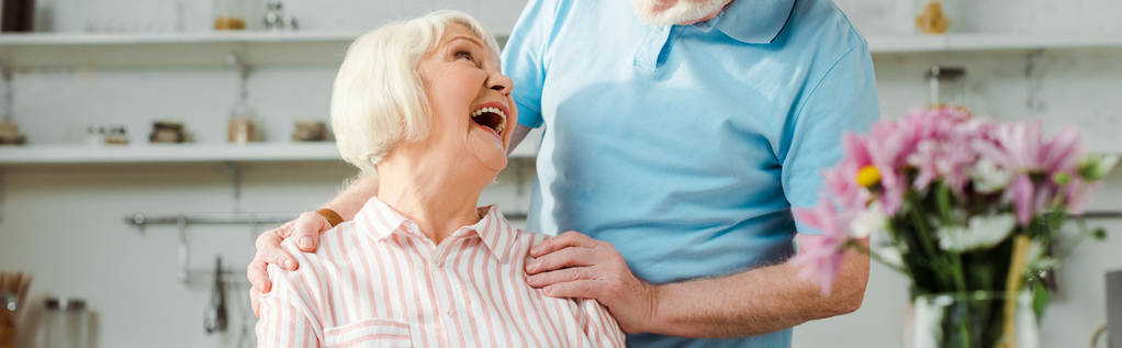 Πανοραμική φωτογραφία της ηλικιωμένης γυναίκας να γελάει και να κοιτάζει τον άντρα της στην κουζίνα  - Φωτογραφία, εικόνα