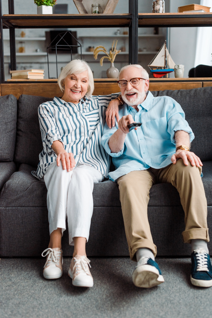 Lächelndes Seniorenpaar mit Fernbedienung sitzt auf Sofa im Wohnzimmer - Foto, Bild