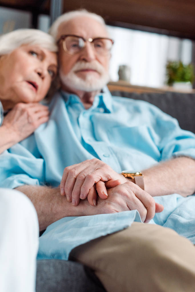 Επιλεκτική εστίαση του ηλικιωμένου ζευγαριού κρατώντας τα χέρια, ενώ κάθεται στον καναπέ στο σπίτι - Φωτογραφία, εικόνα