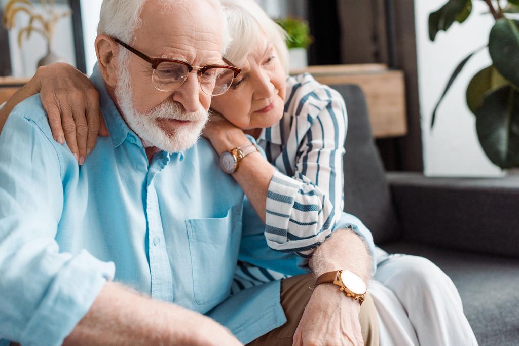 Seniorin umarmt traurigen Ehemann auf Couch zu Hause - Foto, Bild