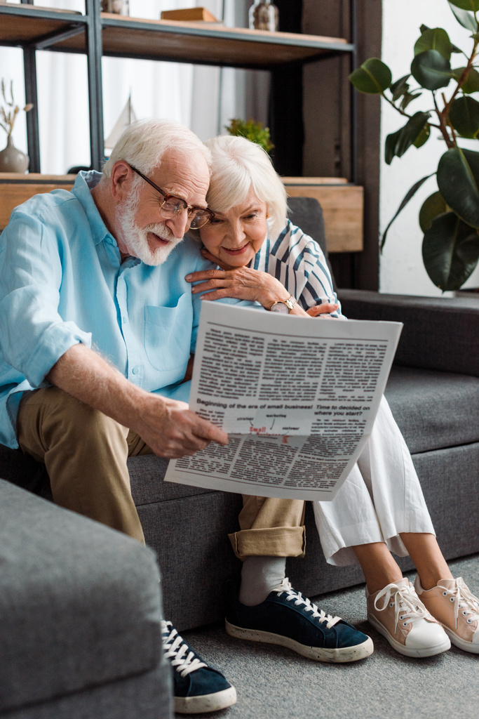 Mujer mayor sonriente sentada por el marido con el periódico en el sofá en casa
 - Foto, Imagen