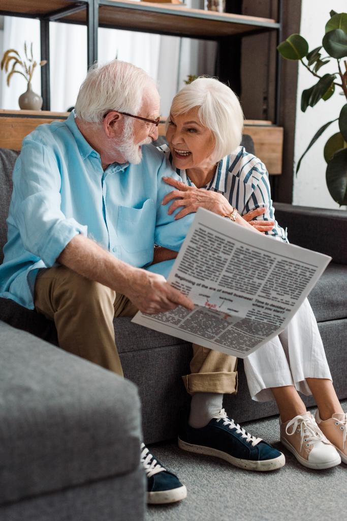 Starsza para uśmiechająca się do siebie i mężczyzna trzymający gazetę na kanapie w domu - Zdjęcie, obraz