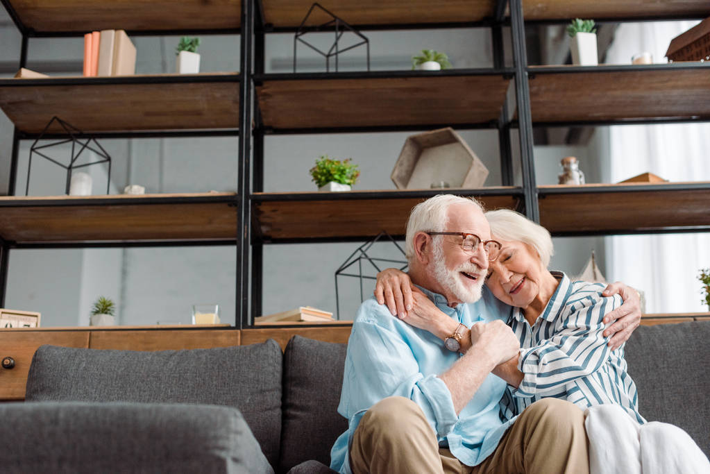 Low angle view of smiling senior man embracing wife on couch in living room - Photo, Image