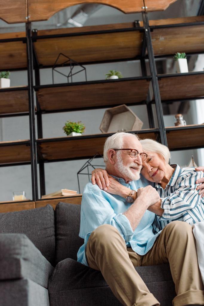 Low angle view of smiling senior couple hugging on couch at home - Photo, Image