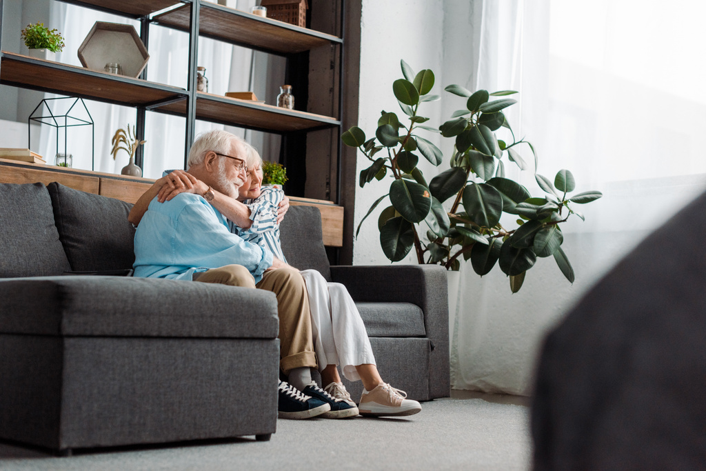 Вид сбоку пожилой пары, обнимающейся на диване в гостиной
 - Фото, изображение