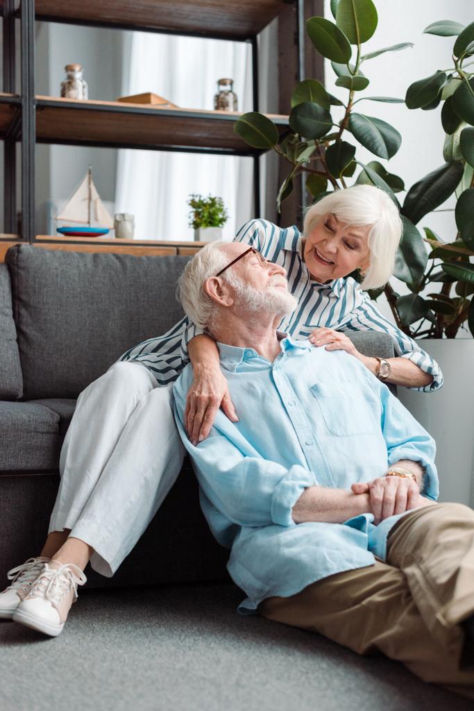 glimlachende senior vrouw knuffelen echtgenoot op vloer in woonkamer - Foto, afbeelding
