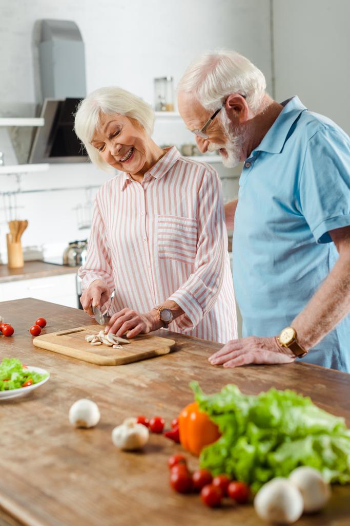 Выборочный фокус улыбающегося мужчины, стоящего рядом с женой, обрезающего свежие овощи на кухонном столе
 - Фото, изображение