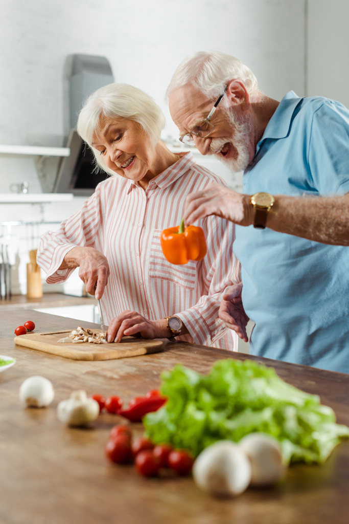 Selektivní zaměření seniora drží paprika tím, že se usmívá manželka krájení hub v kuchyni - Fotografie, Obrázek