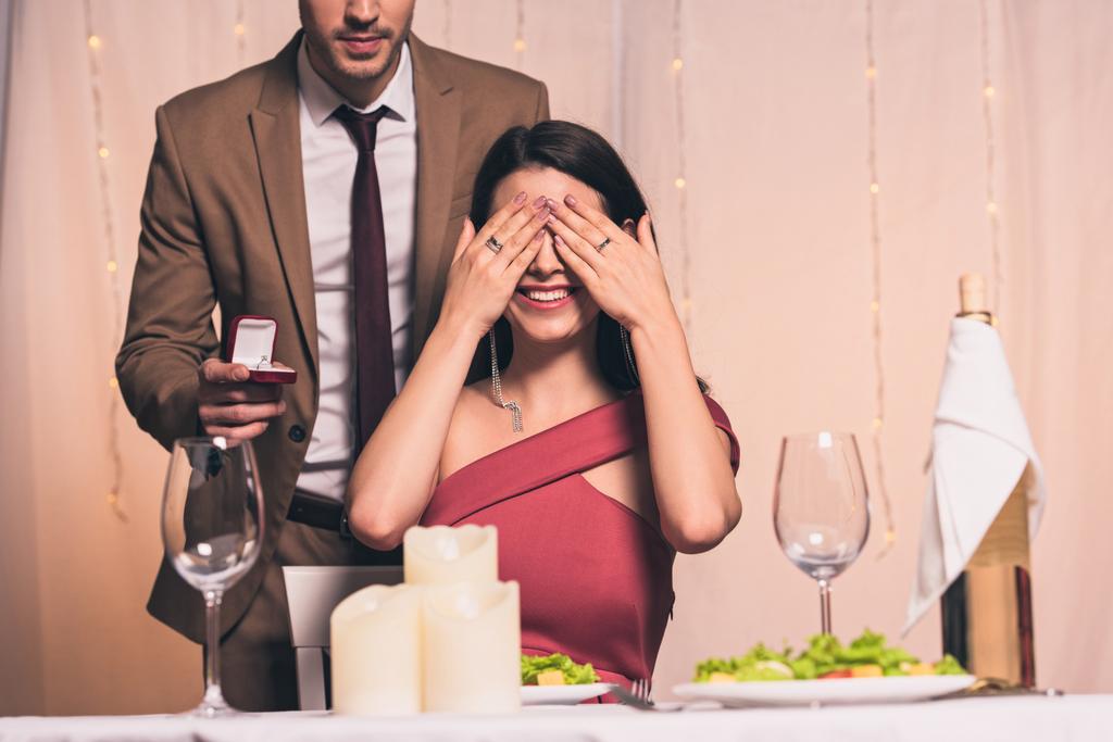 елегантний чоловік тримає обручку біля щасливої дівчини, сидячи за столом і прикриваючи очі руками
 - Фото, зображення
