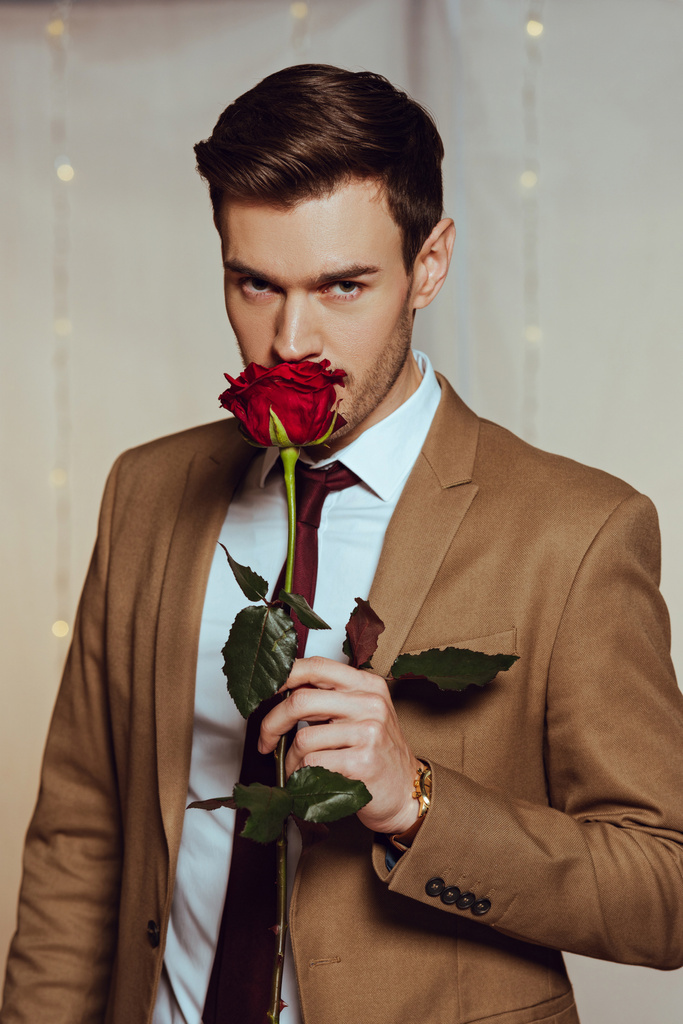 handsome, elegant man sniffing red rose while standing in restaurant and looking at camera - Photo, Image