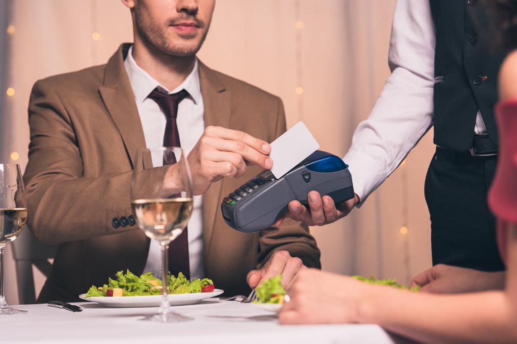 vista recortada de un hombre elegante que paga con tarjeta de crédito con terminal mientras está sentado en el restaurante cerca de la novia
 - Foto, imagen