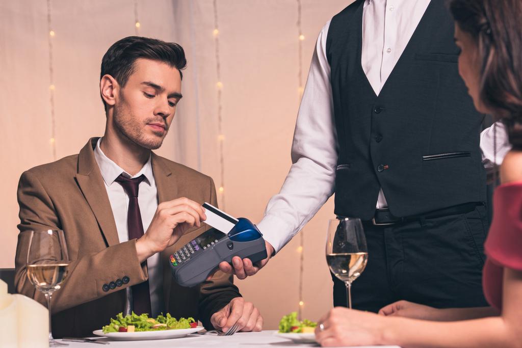 schöner, eleganter Mann bezahlt mit Kreditkarte mit Terminal, während er im Restaurant in der Nähe seiner Freundin sitzt - Foto, Bild