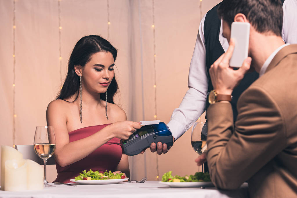 елегантна дівчина платить кредитною карткою з терміналом в ресторані, поки хлопець розмовляє на смартфоні
 - Фото, зображення