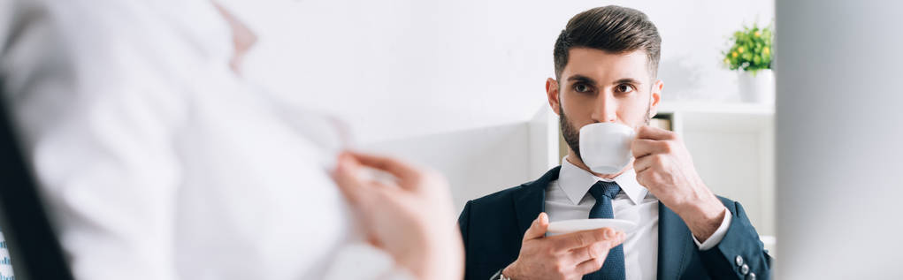 πανοραμική φωτογραφία του επιχειρηματία πίνοντας καφέ και κοιτάζοντας γραμματέα στο γραφείο  - Φωτογραφία, εικόνα