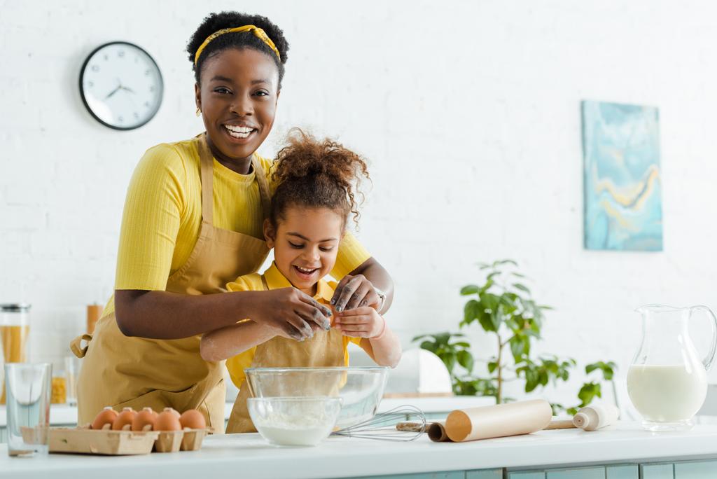 かわいいアフリカ系アメリカ人の子供と陽気な母親が台所で調理中にボウルに卵を追加  - 写真・画像
