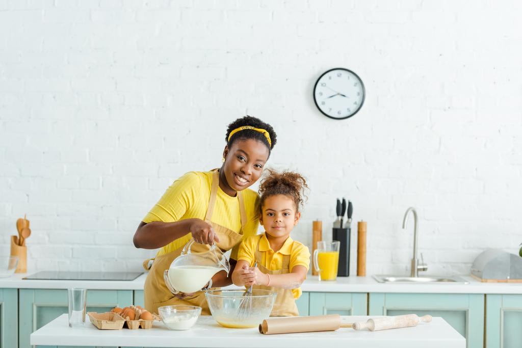 cheerful african american mother pouring milk in bowl near cute daughter and looking at camera - Photo, Image