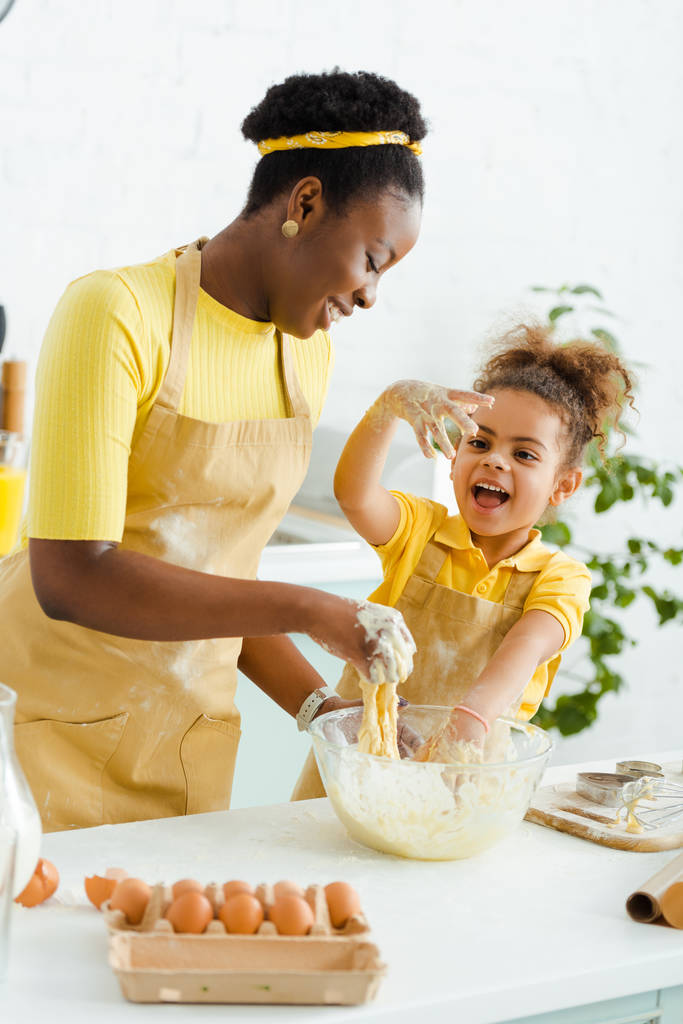szczęśliwy Afryki amerykański matka ugniatając ciasto w pobliżu wesoły córka  - Zdjęcie, obraz