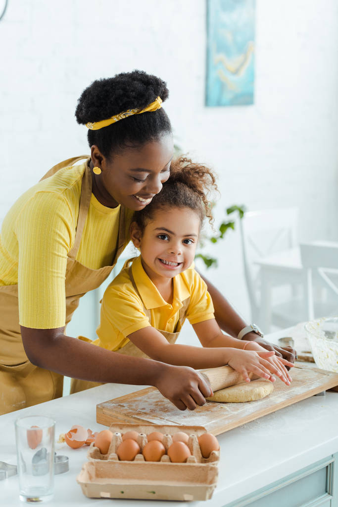 attrayante mère afro-américaine regardant la pâte crue près de fille mignonne et tenant rouleau à pâtisserie
  - Photo, image
