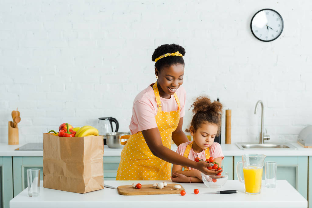Mutlu Afro-Amerikan annesi kızının yanındaki kaseye kiraz domates koyuyor.  - Fotoğraf, Görsel