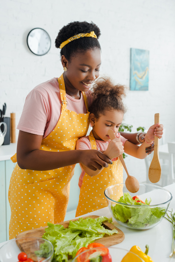 alegre africano americano madre y lindo niño celebración de madera cuchara y espátula mientras preparar ensalada
  - Foto, Imagen
