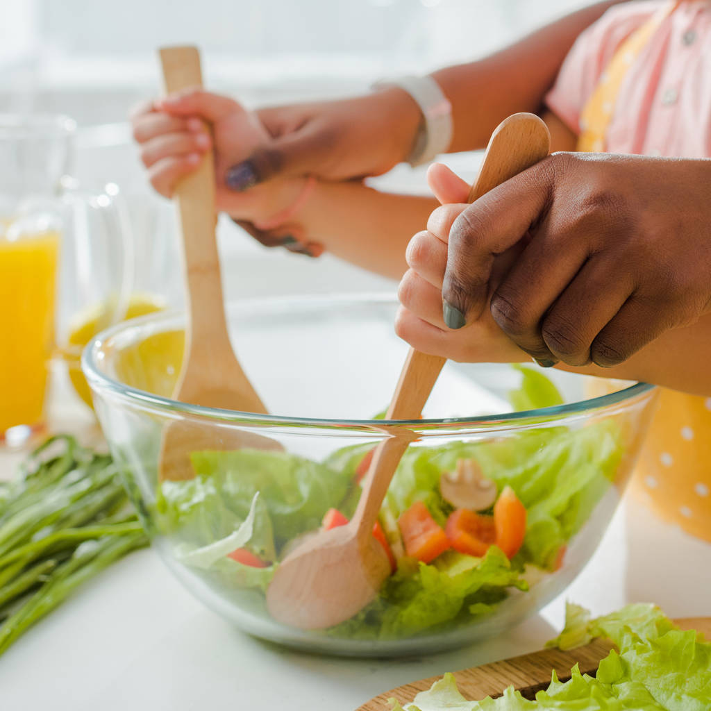 abgeschnittene Ansicht einer afrikanisch-amerikanischen Mutter mit Kind, die Kochlöffel und Spachtel hält, während sie Salat mixt  - Foto, Bild