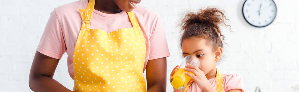 Панорамный снимок счастливой африканской матери-американки, улыбающейся рядом с милой дочерью, пьющей свежий апельсиновый сок
  - Фото, изображение
