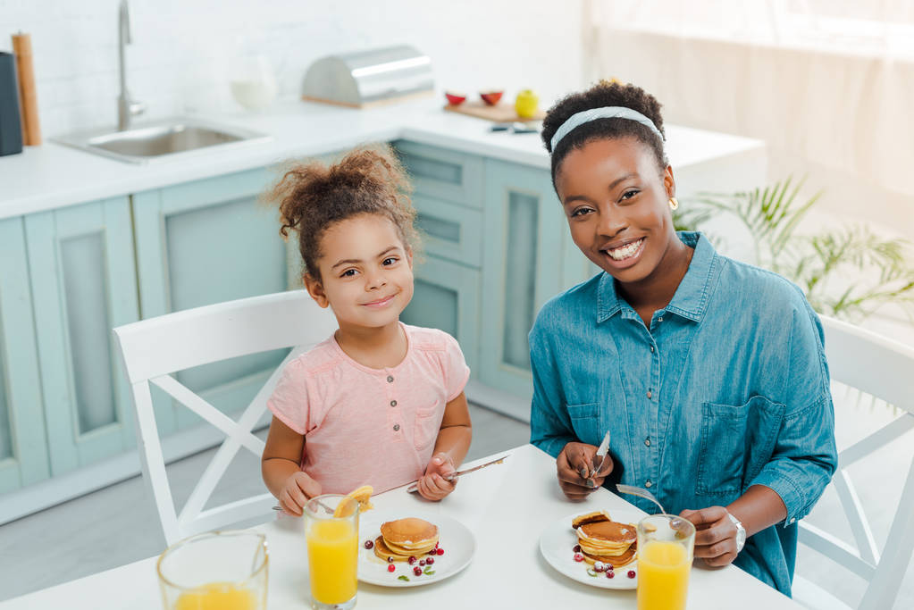 happy african american mother and daughter looking at camera near tasty pancakes  - Photo, Image