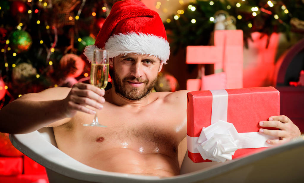 Zdrówko. Świąteczny prezent. Seksowny brodaty Mikołaj relaksujący się w gorącej kąpieli z szampanem i pudełkiem prezentów. Człowiek muskularny tułów leżących w wannie dekoracje świąteczne tle. Wesołych Świąt koncepcja - Zdjęcie, obraz