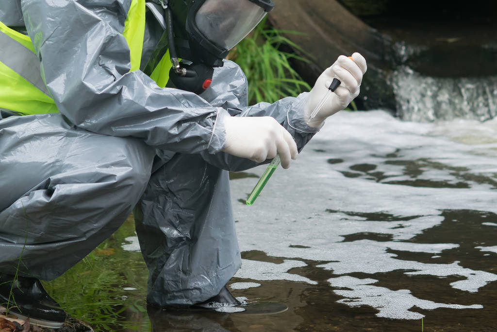 un hombre con traje protector y máscara sostiene un tubo de ensayo y una pipeta con agua de prueba en el río después de un accidente
 - Foto, Imagen