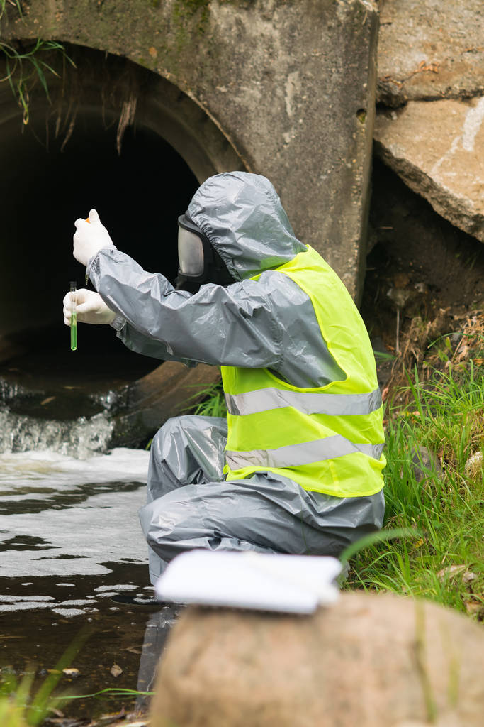 boční pohled na muže v ochranném obleku a masce držícího zkumavku a pipetu s testovací vodou na řece po nehodě venku - Fotografie, Obrázek