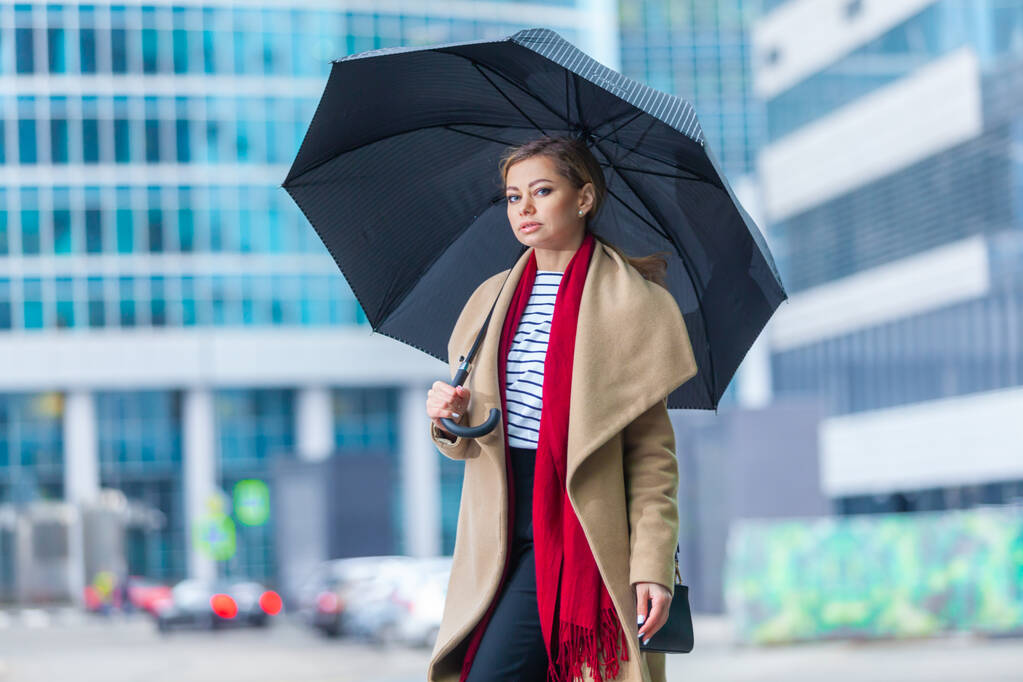 Regen. Outdoor Lifestyle Mode Porträt von atemberaubenden brünetten Mädchen. zu Fuß auf der Stadtstraße. Einkaufen gehen. Er trug einen schicken weißen Mantel, ein rotes Halstuch und einen schwarzen Regenschirm. Geschäftsfrau. - Foto, Bild