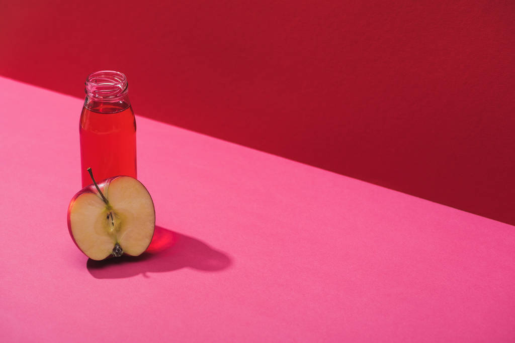 свежий сок в бутылке рядом с яблоком половина на красном и розовом фоне
 - Фото, изображение