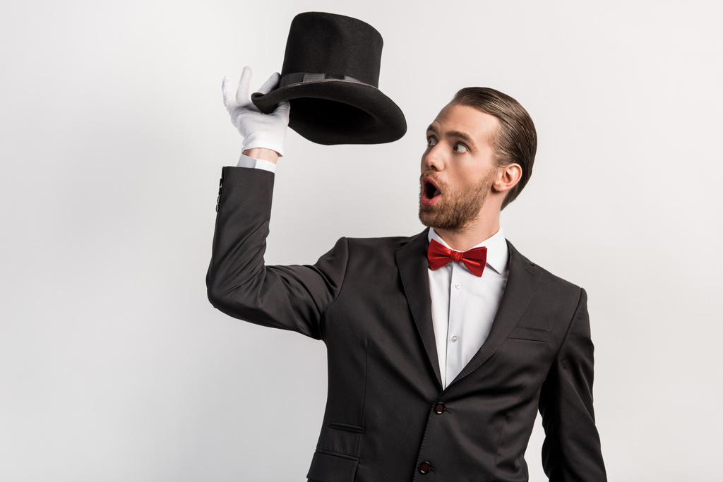 shocked magician with open mouth looking at hat, isolated on grey - Photo, Image