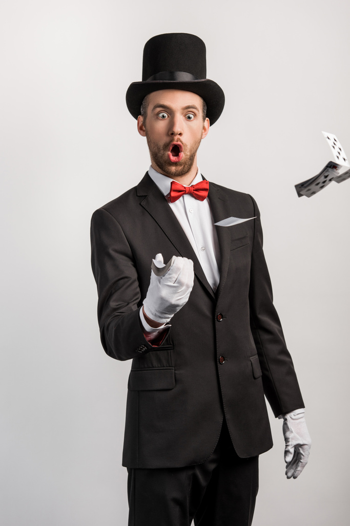 šokovaný kouzelník s otevřenými ústy házet hrací karty, izolované na šedé - Fotografie, Obrázek