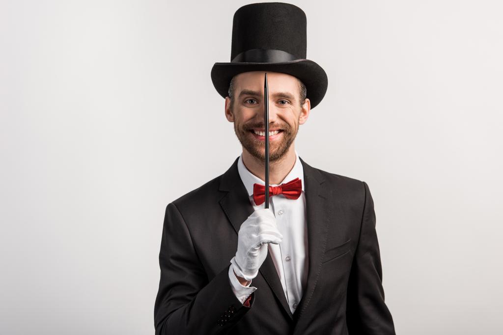veselý kouzelník v obleku a klobouku držící hůlku, izolovaný na šedi - Fotografie, Obrázek