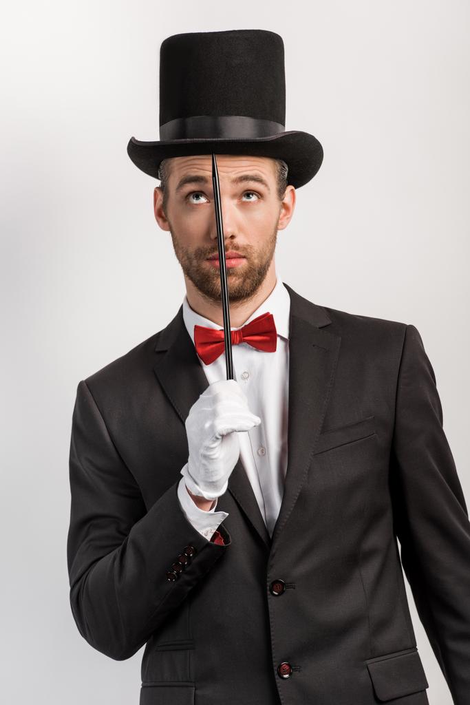 přemýšlivý kouzelník v obleku a klobouku držící hůlku, izolovaný na šedi - Fotografie, Obrázek