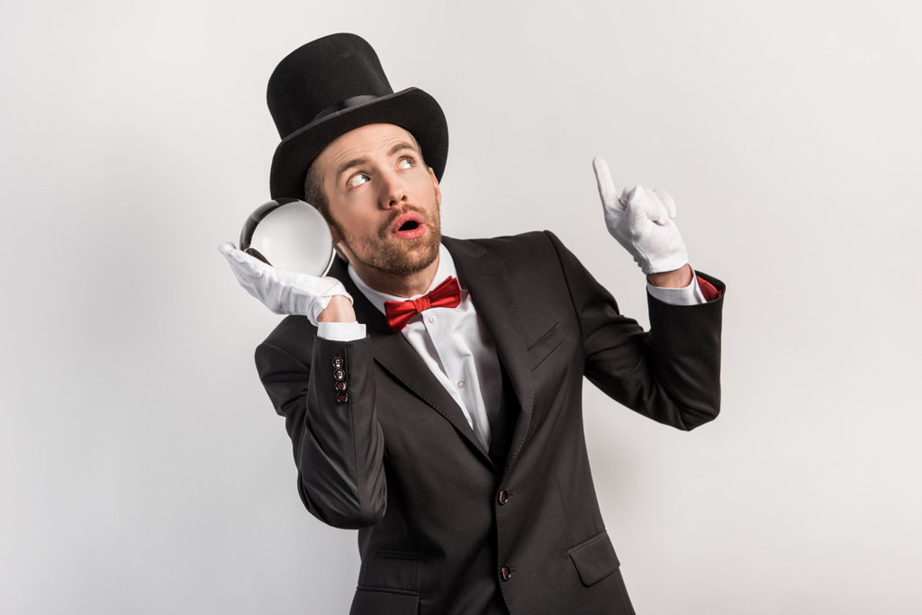 professional magician pointing up and listening to magic ball, isolated on grey - Photo, Image