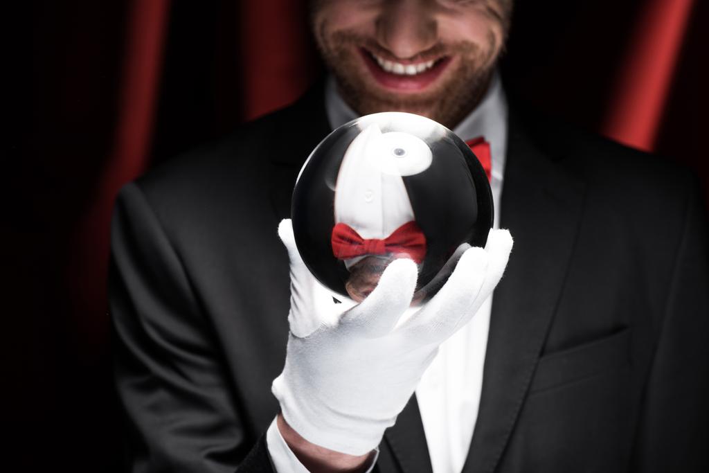oříznutý pohled na usmívajícího se kouzelníka držícího kouzelný míč v cirkusu s červenými závěsy - Fotografie, Obrázek