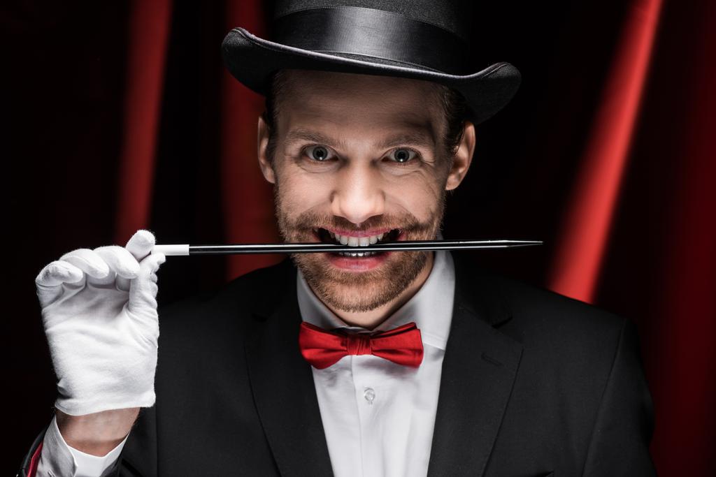 professionele enge goochelaar in pak en hoed stok in tanden in circus met rode gordijnen - Foto, afbeelding
