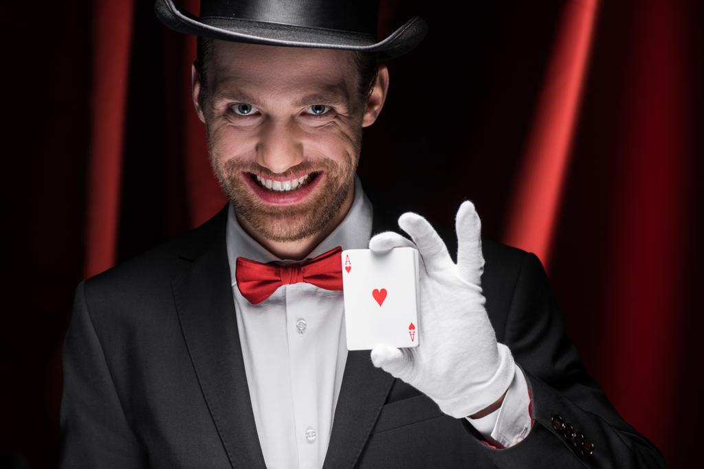 glimlachende goochelaar houdt speelkaarten in circus met rode gordijnen - Foto, afbeelding