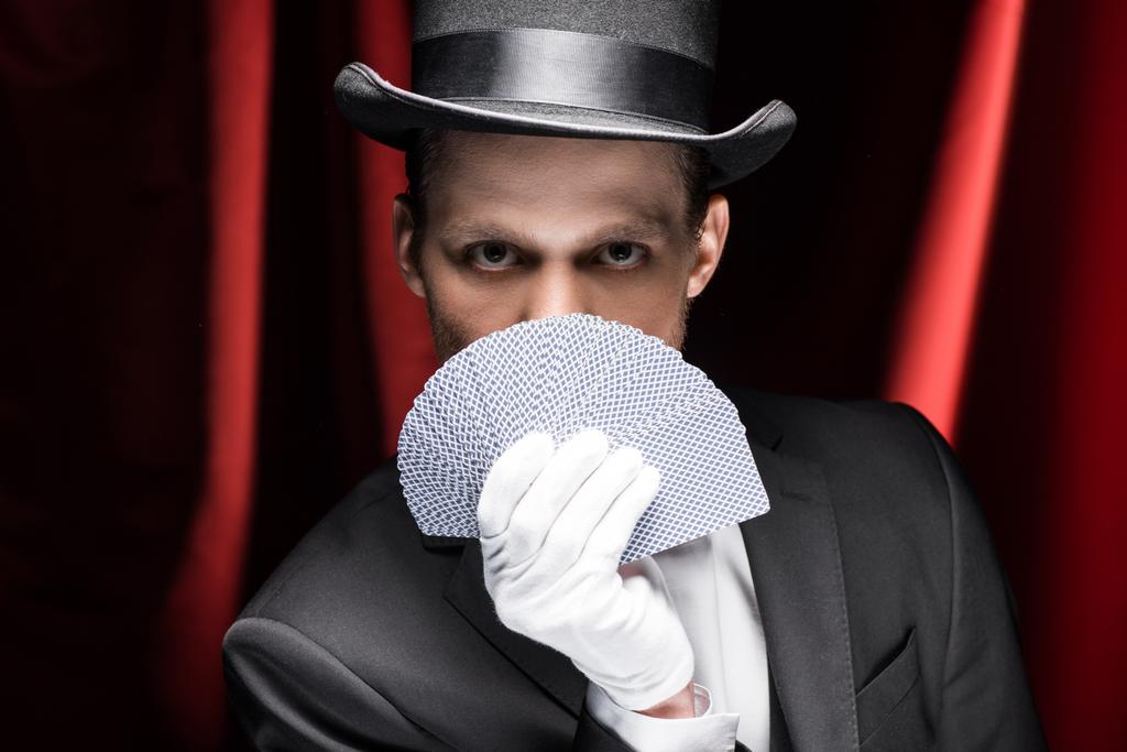 mágico em chapéu segurando cartas de jogo no circo com cortinas vermelhas
 - Foto, Imagem