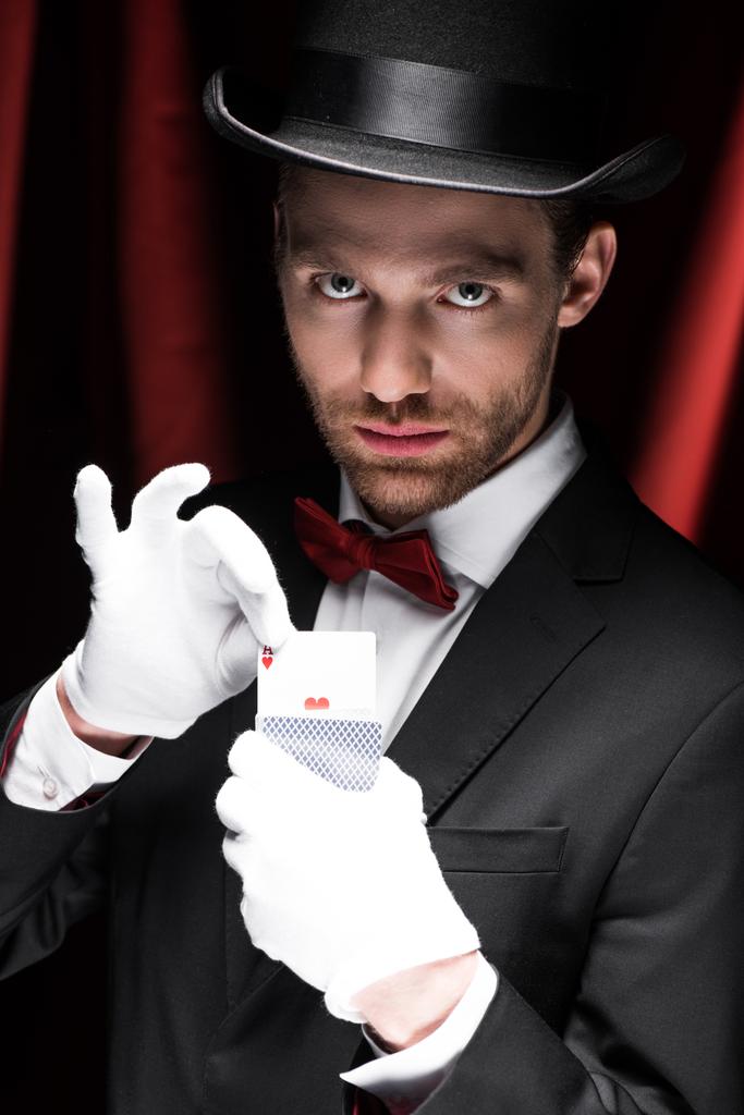 seriózní kouzelník drží hrací karty v cirkusu s červenými závěsy - Fotografie, Obrázek