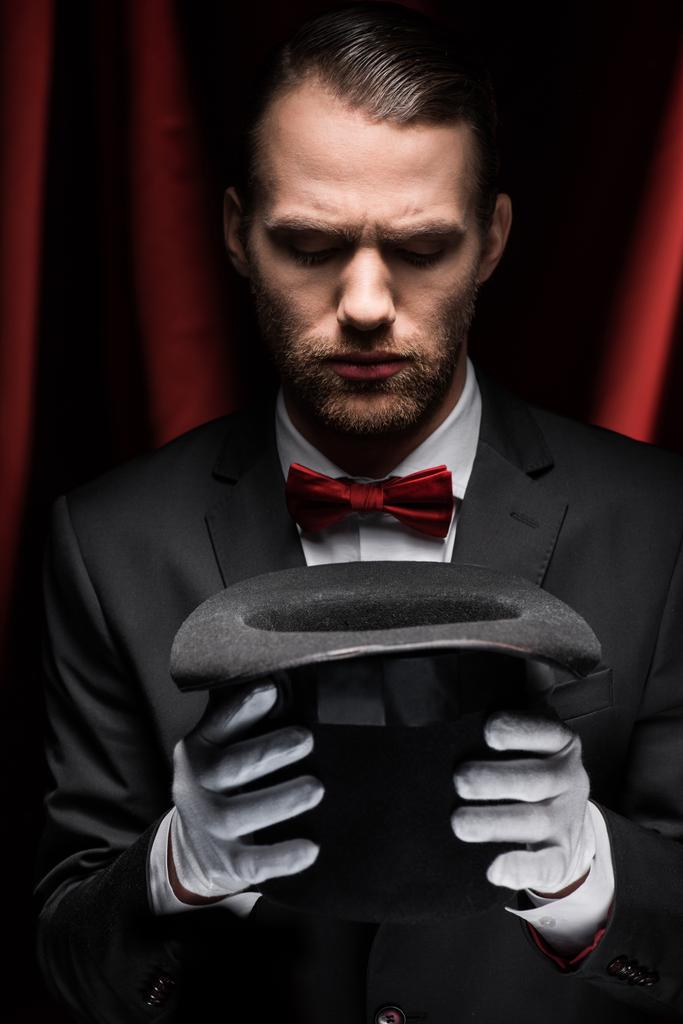Zauberer im Anzug blickt auf Hut im Zirkus mit roten Vorhängen - Foto, Bild