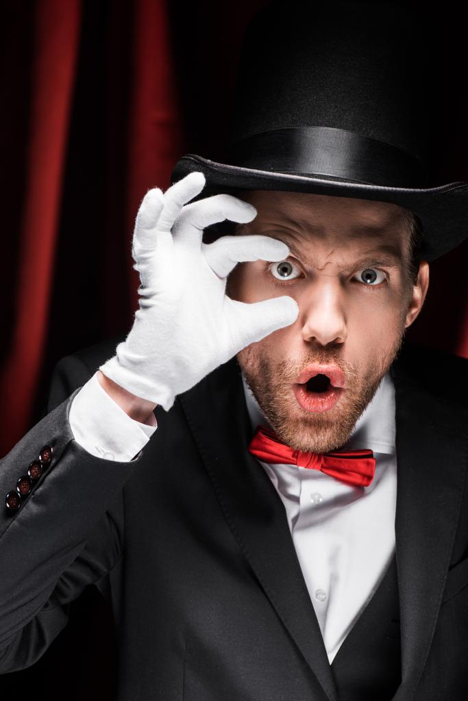 shocked magician opening eye with fingers in circus with red curtains - Photo, Image
