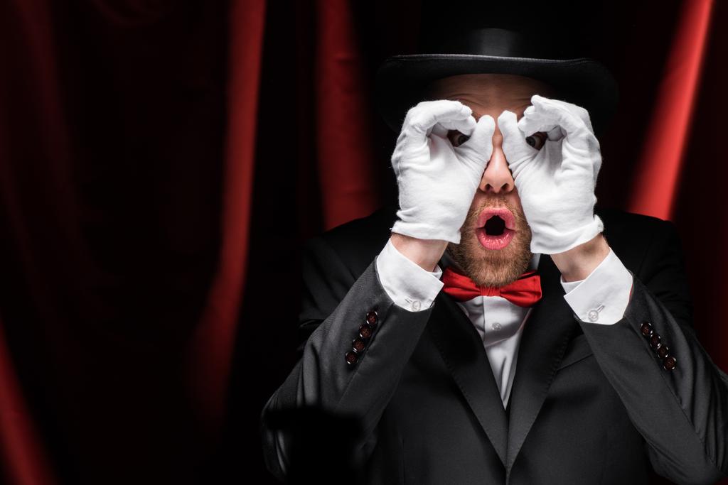 wstrząśnięty magik robi okulary z rąk w cyrku z czerwonymi zasłonami - Zdjęcie, obraz