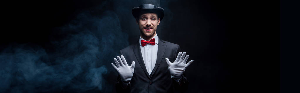 panoramisch schot van gelukkige goochelaar in pak en hoed gebaren in donkere rokerige kamer - Foto, afbeelding