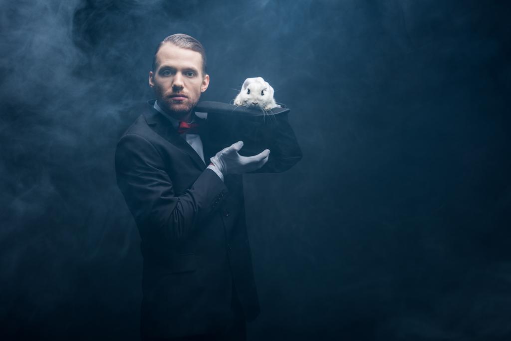 Takım elbiseli profesyonel sihirbaz şapkalı beyaz tavşanla numara yapıyor, dumanlı karanlık oda - Fotoğraf, Görsel